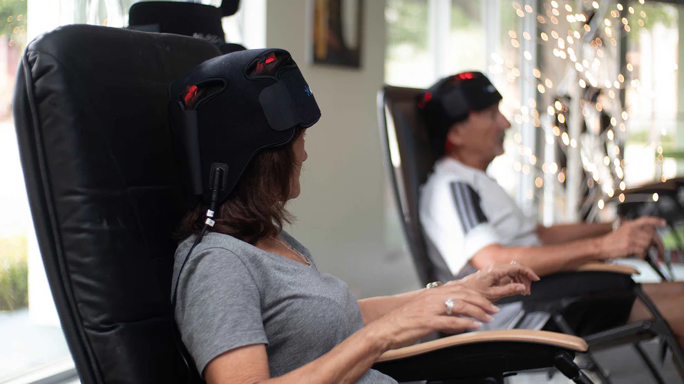 Two people using black gaming chairs in a home setting with decorative lights in the background.