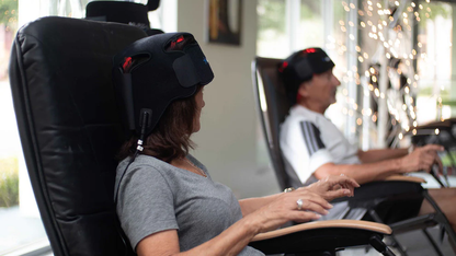Two people using black gaming chairs in a home setting with decorative lights in the background.